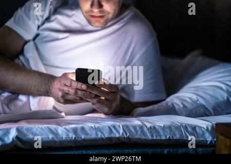 Man in bed with phone at night. Texting with smartphone before sleeping. Guy holding cellphone in hand. Dark home bedroom. Infidelity, cheating. Stock Photo