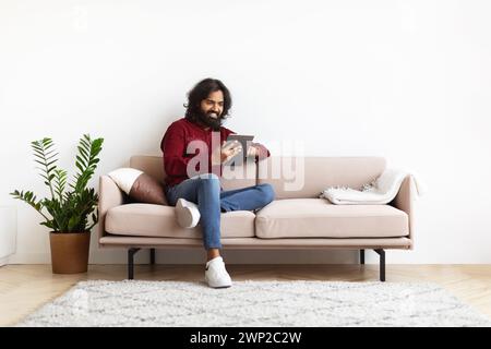 Cheerful young indian man sitting on couch, using digital tablet Stock Photo