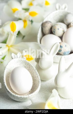 Holiday Easter composition with eggs in marble bowls with bunny ears, small ceramic bunnies, white yellow Iris flowers on white tablecloth. Festive wh Stock Photo