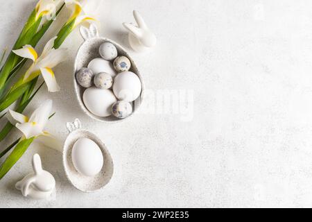 Trendy monochrome white Easter card with natural colors eggs in marble bowls with bunny ears, small ceramic bunnies, bouquet of white Iris flowers on Stock Photo