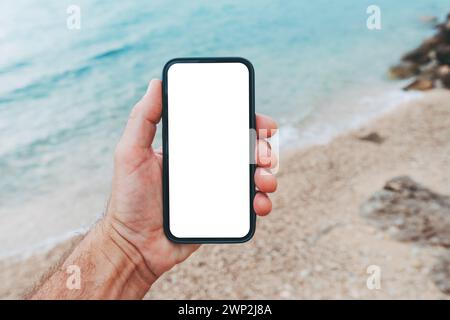 Smartphone mockup at the ocean beach, closeup of male hand holding mobile phone with blank white screen as copy space at seaside coast in summer Stock Photo