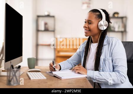 Smiling black female student with headphones taking notes from online lecture Stock Photo