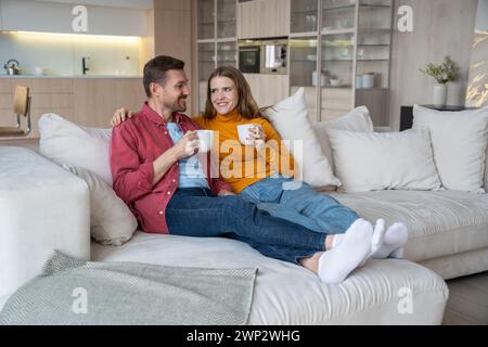 Newlyweds hugging lie on huge home sofa. Young couple happily drinks coffee enjoying weekend time. Stock Photo