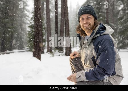 A man wearing a blue and gray jacket and a black hat is smiling in the snow. He is sitting on the ground and he is enjoying the winter weather Stock Photo