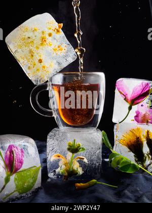 a transparent cup of tea in ice, ice in the form of frozen cubes with flowers, hot tea is pouring, steam is coming from the tea, the background is dar Stock Photo