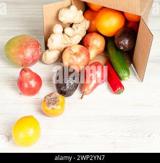 Delivery of fruits and vegetables in a cardboard box. The box is open on a wooden table, rolled out of it - lemon, persimmon, pears, avocado hass, man Stock Photo