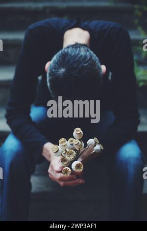 A woman with bowed head and short shaved hair, sitting on a step holding a bouquet of dried poppy flowers. Stock Photo