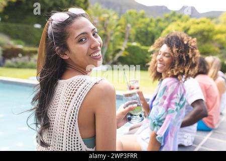 Young biracial woman smiles at the camera, friends enjoy drinks by the pool Stock Photo