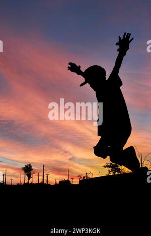 silhouette of skater boy with beanie jumping in profile in the air with his hands up in the air with a sunset in the background Stock Photo