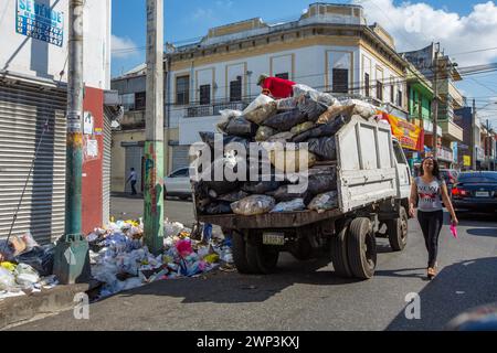 Men load garbage off the sidewalk onto a truck in Santo Domingo, Dominican Republic as a young woman walks by. Stock Photo