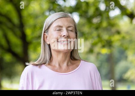 Close-up portrait of smiling senior gray-haired woman in pink t-shirt standing in park and resting, breathing relaxed with eyes closed after walking and doing sports. Stock Photo