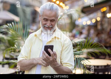 Close-up photo of an older gray-haired man in a yellow shirt standing on a city street and using a mobile phone. Stock Photo