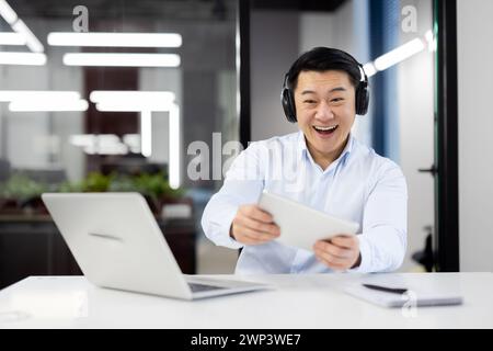 Smiling young man working in the office, sitting at a desk with a laptop, wearing headphones and playing online games on a tablet during a break. Stock Photo