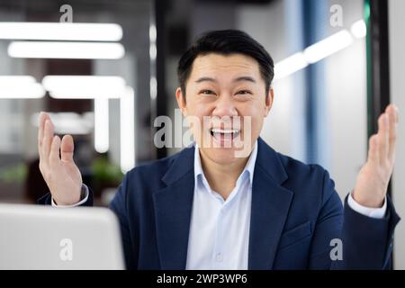 Close-up portrait of a happy and successful young Asian businessman man in a suit sitting in the office at a table in front of a laptop, smiling and looking at the camera with his arms outstretched. Stock Photo