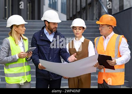 Focused diverse group of engineers with hard hats discussing blueprints in front of an office building. Stock Photo