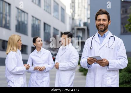 Smiling male doctor with tablet standing in front of hospital, engaged in conversation with a team of healthcare workers. Stock Photo