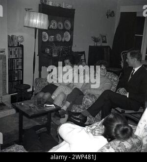 1960s, historical, a family sitting together in a living room, England, UK. In the room, traditional furniture, small bookcase, standing lamp and dresser with plates. No mobile phones on laps, but typically for the era, a lady sitting on the sofa beside an ashtray and packet of cigarettes. Stock Photo