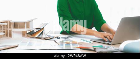 Cropped image of professional interior designer using laptop to create house plan on table with color palette and architectural model scatter around Stock Photo