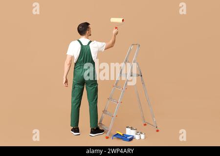 Portrait of male painter with paint roller and stepladder painting something on beige background Stock Photo