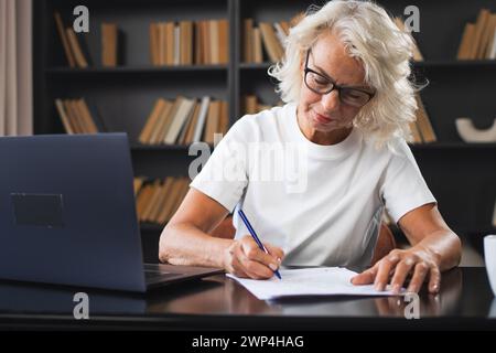 Middle aged senior woman using laptop computer writing notes at workplace. Focused mature old businesswoman work at office making written records doing online work. Mature boss professional worker Stock Photo
