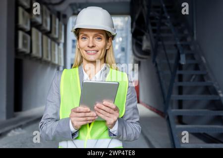 Professional caucasian woman in helmet and neon yellow vest standing at construction with digital tablet in hands. Experienced female architect checking house building process and entering data. Stock Photo