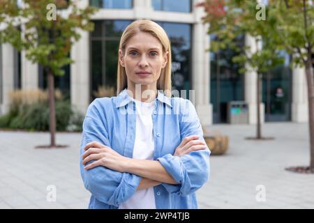 Serious blonde woman in blue shirt standing alone near glass corporate building with hands folded on chest. Successful business owner feeling confident in career development and opportunities. Stock Photo
