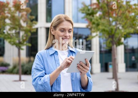 Smiling blonde woman using digital tablet while standing outdoors at pedestrian space with trees. Confident landscape designer developing green zone area in district with futuristic skyscrapers. Stock Photo