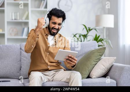 Excited middle eastern person resting on couch with cushions while holding modern tablet and making winner gesture. Joyful male playing online video game in wireless headset at domestic area. Stock Photo