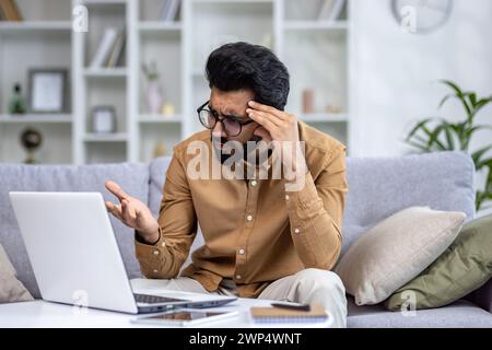 Worried arab male in eyeglasses looking at portable computer screen with confusion while sitting at pillowed sofa. Stressed technical student struggling with learning information from remote class. Stock Photo