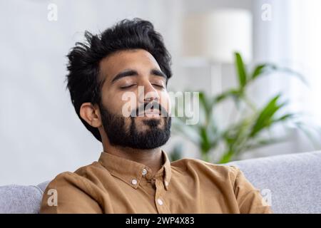 Portrait of indian bearded man with trendy hairstyle leaning on grey sofa back and closing eyes in calmness. Relaxed guy daydreaming and trying taking nap on background of living room interior. Stock Photo
