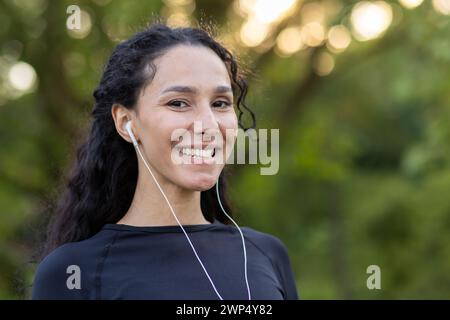 Optimistic diverse sportswoman in black top smiling at camera while listening to motivating playlist in wired earphones. Attractive female in black top finishing training in green public area. Stock Photo