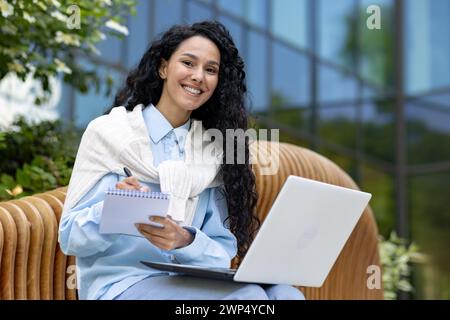 Successful diverse entrepreneur sitting on wooden bench and making notes in daybook while holding computer on laps. Smiling female writing to do list by hand for not skipping important tasks. Stock Photo