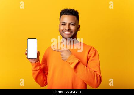 Surprised Indian man holding smartphone in hand and points finger at empty phone screen, astonishment guy presenting best deal, advertising new mobile app, isolated on yellow Stock Photo