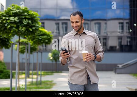 Relaxed middle eastern male having walk by corporate building exterior with plastic coffee cup and cellphone. Happy formal manager enjoying break with hot beverage from work and warm weather outdoors. Stock Photo