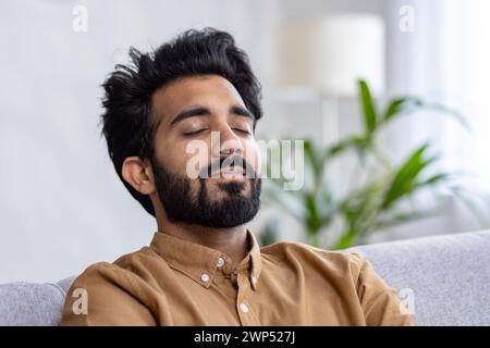 Portrait of indian bearded man with trendy hairstyle leaning on grey sofa back and closing eyes in calmness. Relaxed guy daydreaming and trying taking nap on background of living room interior. Stock Photo