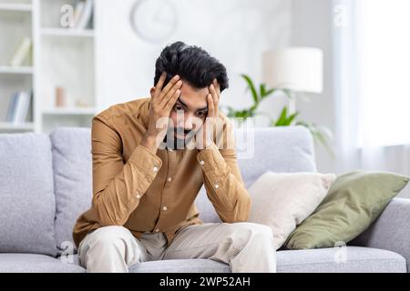 Depressed indian guy holding head with hands while staring at floor and experiencing emotional breakdown. Burnout male struggling with result of overworking while trying focusing and calming down. Stock Photo