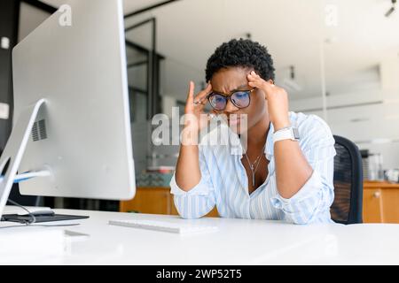 Frustrated african-american businesswoman worrying about project, sitting at the desk in front of PC, depressed female employee feeling stress by mistake or unexpected computer error crash, headache Stock Photo