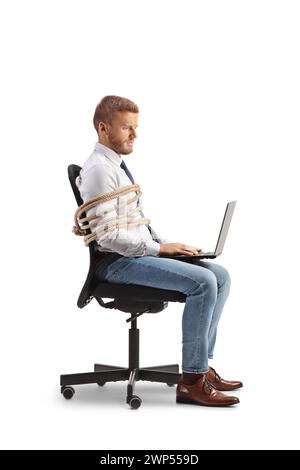 Profile shot of a male office worker sitting in chair tied with a rope and working on a laptop isolated on white background Stock Photo