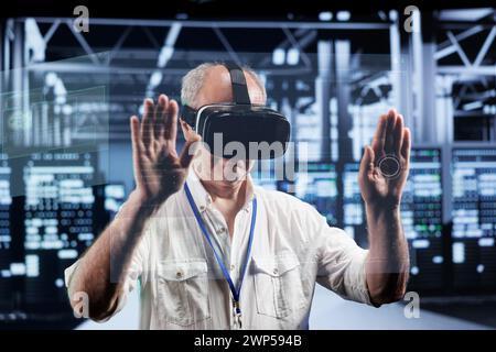 Senescent technical support consultant immersed in virtual reality at data center, doing equipment maintenance. Serviceman using VR headset to optimize server racks performance, checking code lines Stock Photo