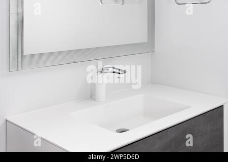 A bathroom faucet detail with a white and chrome faucet, white sink, and a dark grey cabinet. Stock Photo