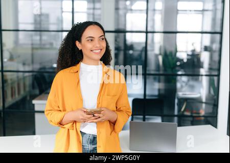 Stylish positive brazilian or hispanic curly haired young woman in an orange shirt, stands at a workplace in the office, holding her smartphone, messaging online, looks away, smiling, dreaming Stock Photo