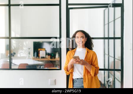 Chatting online. Happy brazilian or hispanic curly haired young woman in casual wear, stands at a modern office space, holding her mobile phone, chatting online, looks at camera, smiling friendly Stock Photo