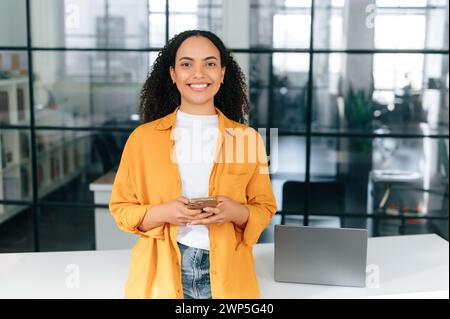 Happy beautiful brazilian or hispanic curly haired young woman in an orange shirt, stands at a workplace in the office, holding her mobile phone, chatting online, looks at camera, smiling friendly Stock Photo