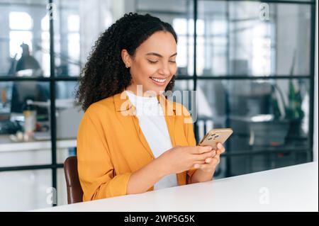 Attractive happy hispanic or brazilian curly haired young woman, office employee, sitting at a workplace, using her smartphone, messaging with colleagues or client, browsing internet, smiling Stock Photo