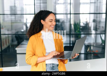 Busy successful positive brazilian or hispanic curly haired woman in casual clothes, company manager, seo, holding an open laptop, standing in a modern creative office, looks at screen, smile Stock Photo