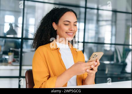 Gorgeous happy hispanic or brazilian curly haired young woman in an orange shirt, sitting at a workplace in the office, using her mobile phone, messaging with friends, browsing social media, smiling Stock Photo