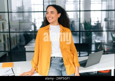 Attractive pretty multiracial curly haired young woman in a casual stylish clothes, stand in the modern office near work desk, looks away, smiling happily, dreaming about something Stock Photo