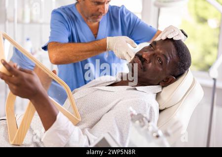 Cosmetologist showing results of facial procedure to dark-skinned man Stock Photo