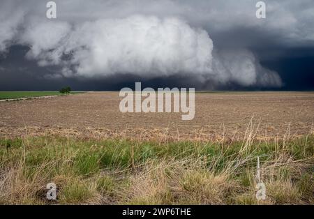 A massive, thick shelf cloud hangs low over flat farmland. A dark storm is approaching fast. Stock Photo