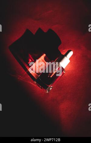 Glass cosmetic pipette bottle on red background top view. Contrasting shadows and decorative cosmetics. Facial serum dark key photo. Fashion magazine. Stock Photo